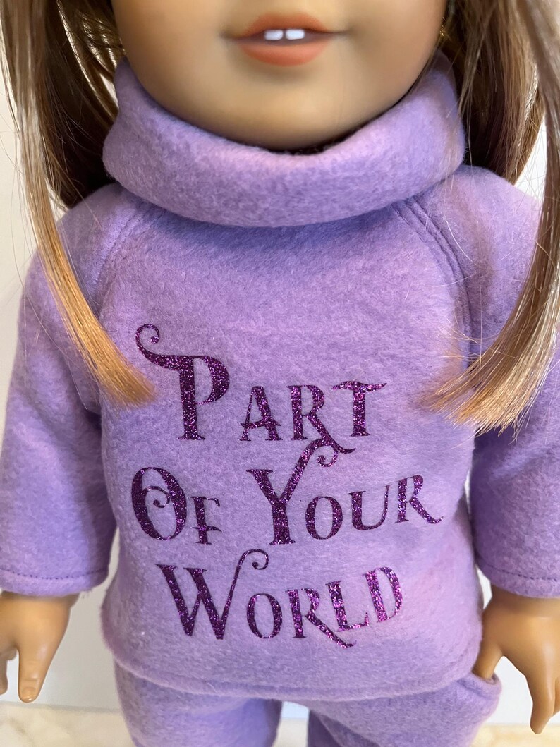 K&ouml;nnte beinhalten: Lilafarbener Fleece-Pyjama f&uuml;r eine Puppe mit dem Text "Part of Your World" in lilafarbenem Glitzer.
