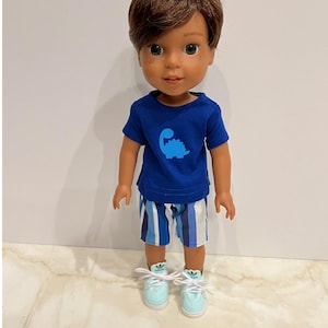 Könnte beinhalten: Eine Puppe trägt ein blaues T-Shirt mit einem blauen Dinosaurier-Motiv, eine blau-weiß gestreifte Shorts und hellblaue Turnschuhe. Die Puppe hat braunes Haar und braune Augen.