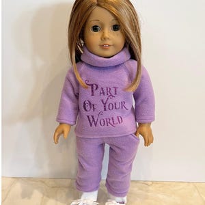 K&ouml;nnte beinhalten: Lilafarbenes Fleece-Outfit f&uuml;r Puppen mit einem Rollkragenpullover und einer passenden Hose. Der Pullover hat den Text "Part of Your World" in lilafarbenem Glitzer.