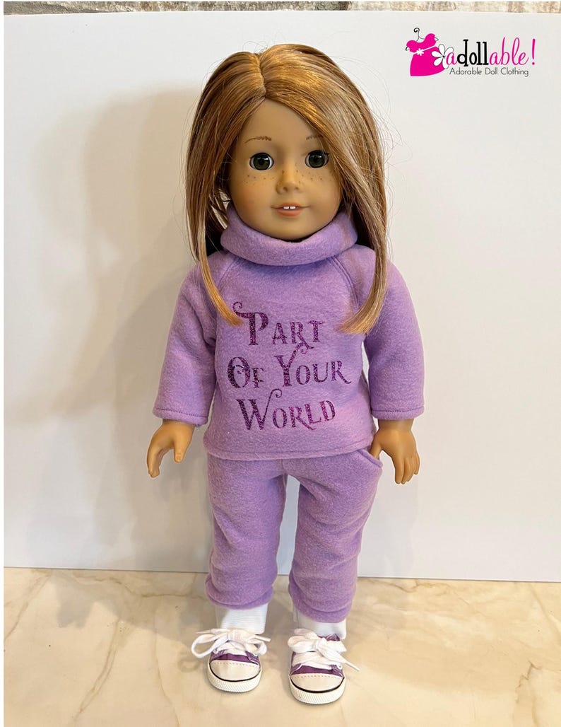 K&ouml;nnte beinhalten: Lilafarbenes Fleece-Outfit f&uuml;r Puppen mit einem Rollkragenpullover und einer passenden Hose. Der Pullover hat den Text "Part of Your World" in lilafarbenem Glitzer.