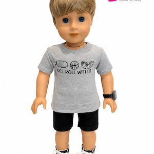 Puede incluir: Una muñeca con una camiseta gris con el texto "JUST ROLL WITH IT" y un gráfico. La muñeca también lleva pantalones cortos negros, zapatillas negras y blancas y un reloj negro. La muñeca tiene el pelo rubio y los ojos azules.