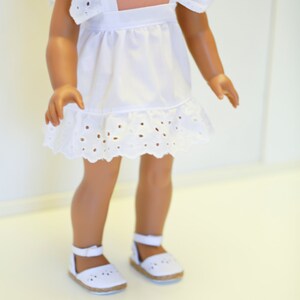 May include: A white eyelet dress with ruffled sleeves and a white eyelet trim. The dress is worn by a doll with brown skin and brown hair. The doll is wearing white sandals.