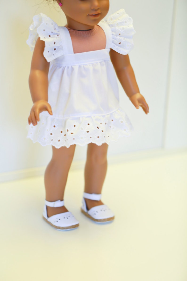 May include: A white eyelet dress with ruffled sleeves and a white eyelet trim. The dress is worn by a doll with brown skin and brown hair. The doll is wearing white sandals.