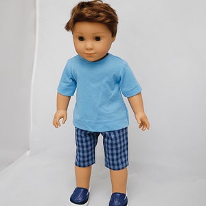 May include: A doll wearing a light blue t-shirt, blue and white checkered shorts, and dark blue sneakers.