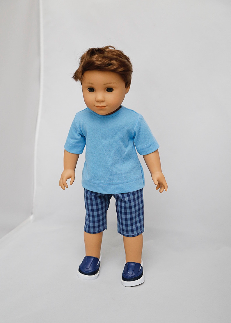 May include: A doll wearing a light blue t-shirt, blue and white checkered shorts, and dark blue sneakers.