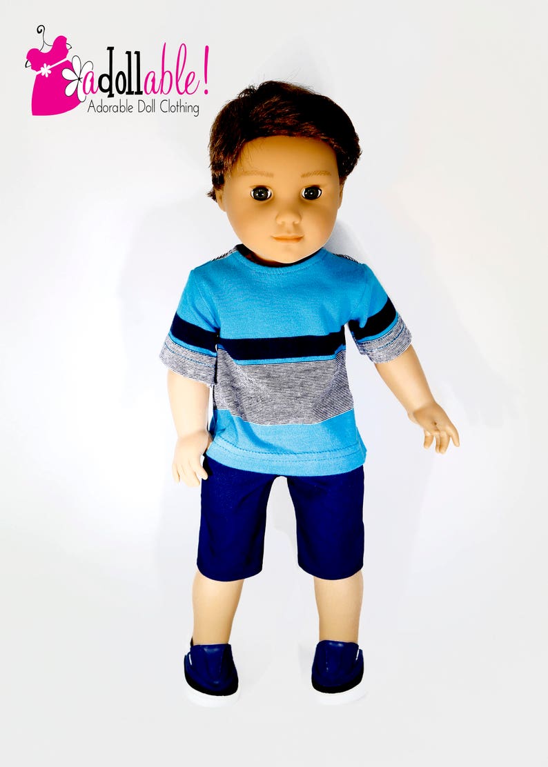 K&ouml;nnte beinhalten: Eine Puppe tr&auml;gt ein blau-graues, gestreiftes T-Shirt mit kurzen &Auml;rmeln, eine dunkelblaue Shorts und dunkelblaue Turnschuhe. Die Puppe hat braunes Haar und braune Augen.