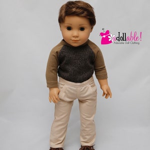 May include: A doll wearing a brown and grey raglan shirt with beige trousers and brown shoes.