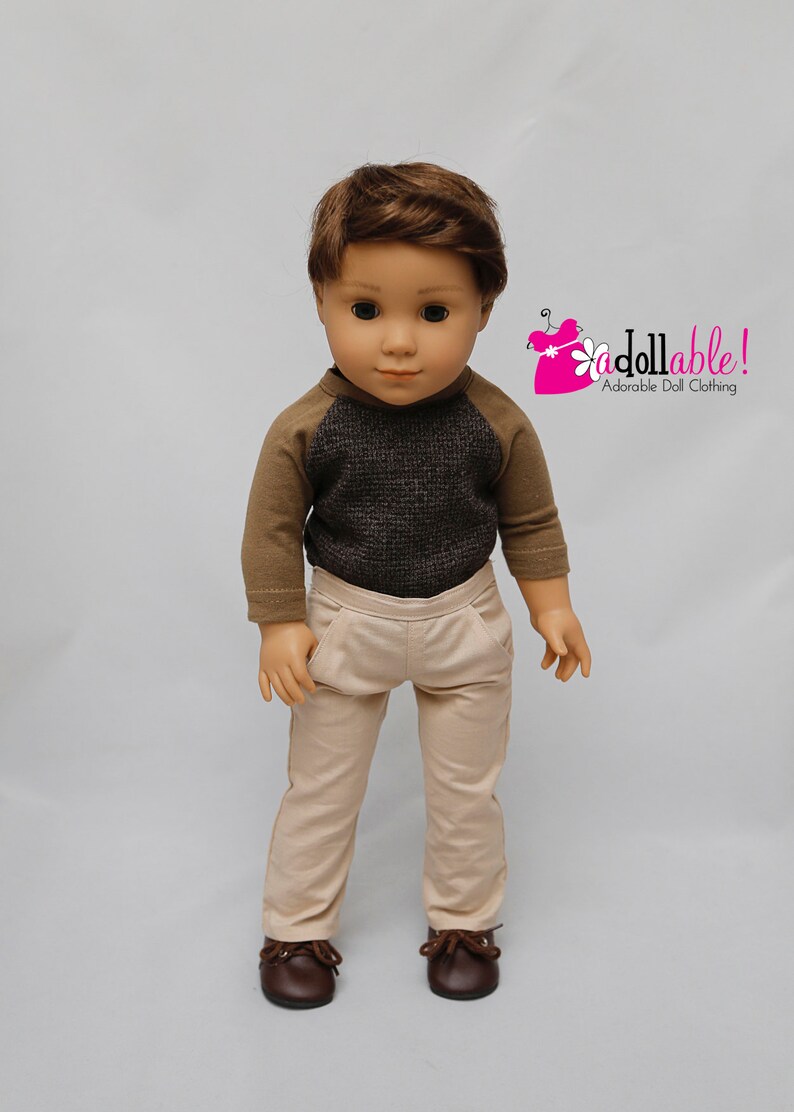 May include: A doll wearing a brown and grey raglan shirt with beige trousers and brown shoes.