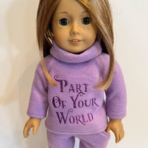 K&ouml;nnte beinhalten: Lilafarbener Fleece-Pyjama f&uuml;r eine Puppe mit dem Text "Part of Your World" in lilafarbenem Glitzer.