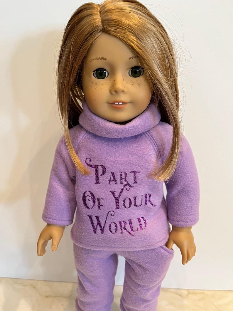 K&ouml;nnte beinhalten: Lilafarbener Fleece-Pyjama f&uuml;r eine Puppe mit dem Text "Part of Your World" in lilafarbenem Glitzer.