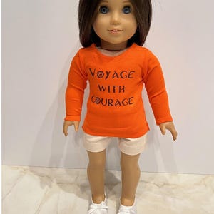May include: Orange long-sleeved shirt with the text "Voyage with Courage" in brown lettering. The shirt is worn by a doll with brown hair and white shoes.