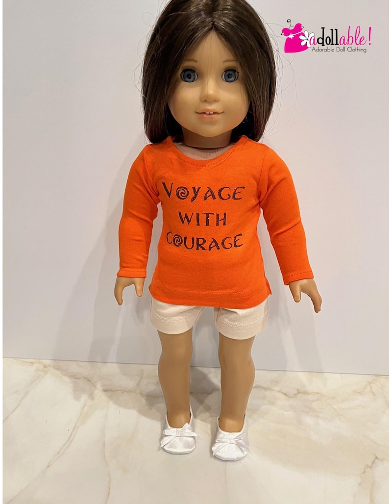 May include: Orange long-sleeved shirt with the text "Voyage with Courage" in brown lettering. The shirt is worn by a doll with brown hair and white shoes.