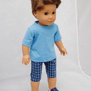 May include: A doll wearing a light blue t-shirt and blue and white checkered shorts. The doll has brown hair and is wearing blue sneakers.
