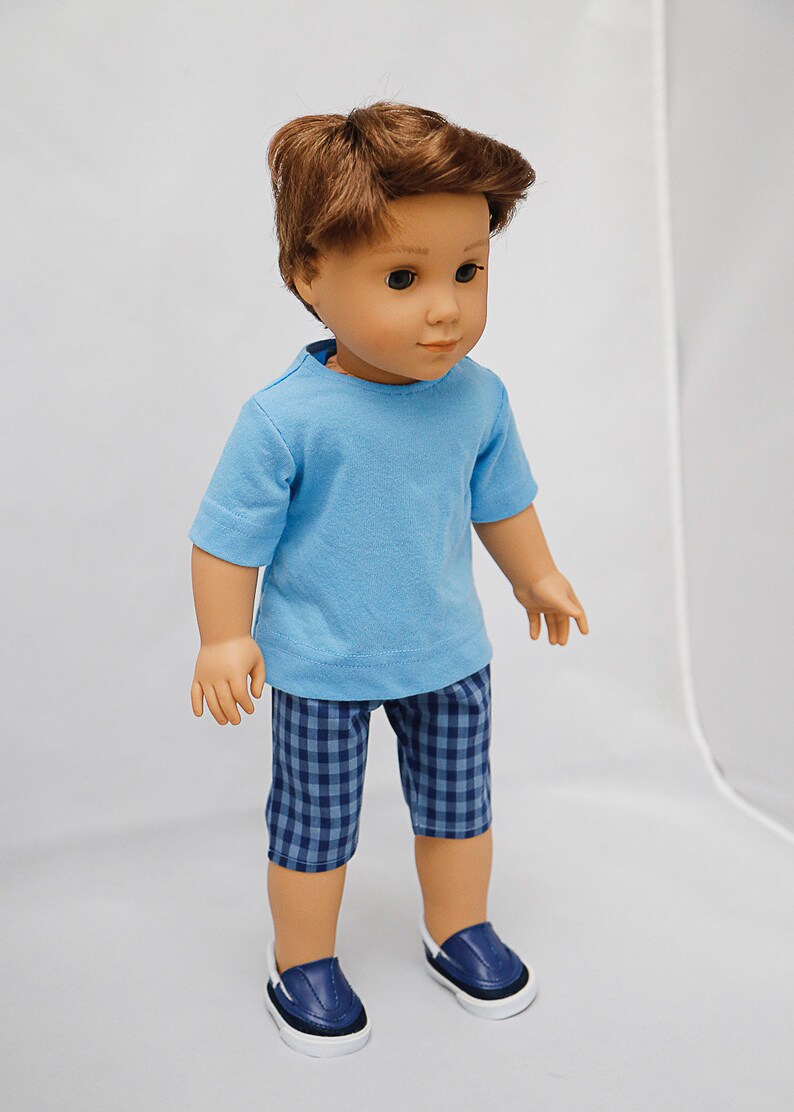 May include: A doll wearing a light blue t-shirt and blue and white checkered shorts. The doll has brown hair and is wearing blue sneakers.