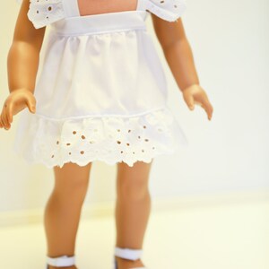 May include: A white dress with eyelet trim and ruffled sleeves. The dress is worn by a doll with brown skin and brown hair. The doll is wearing white shoes.
