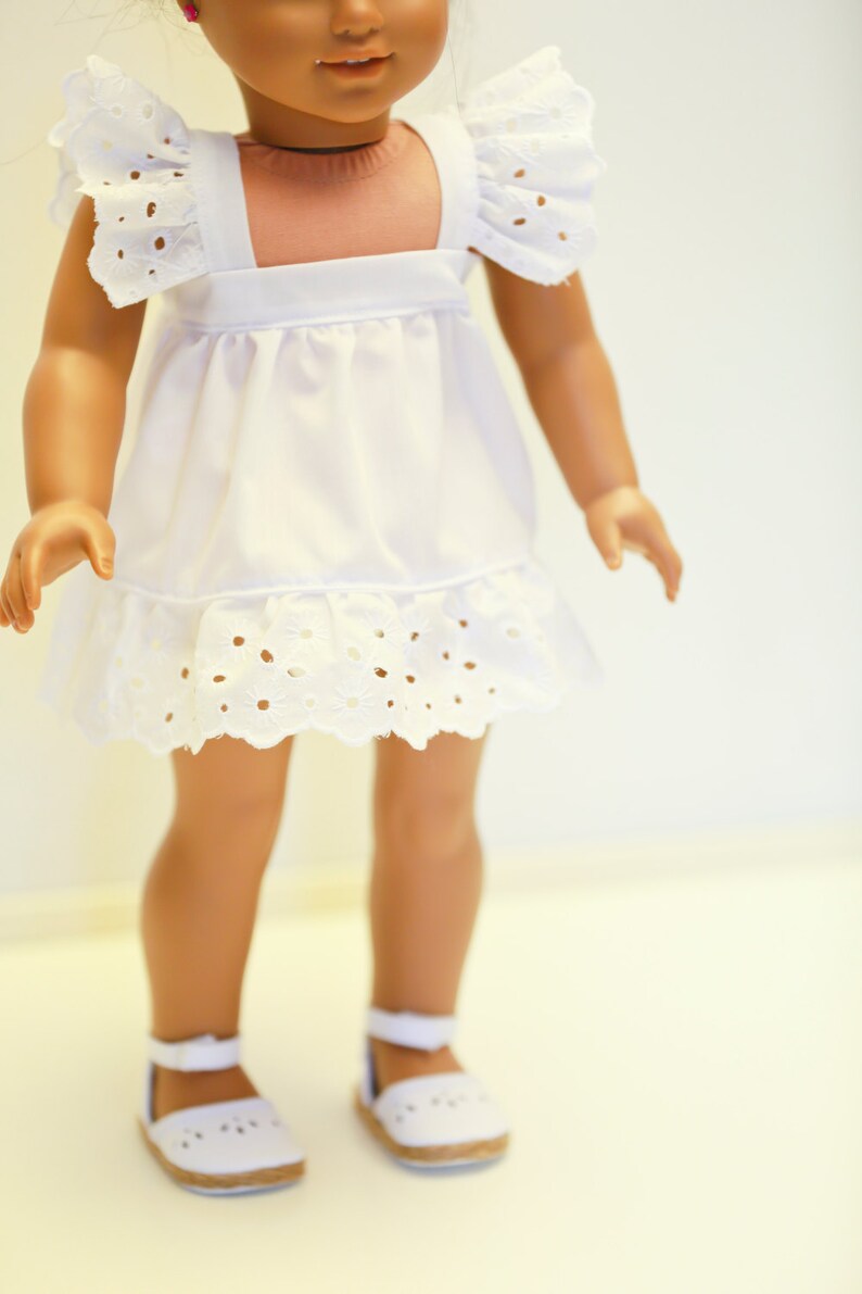 May include: A white dress with eyelet trim and ruffled sleeves. The dress is worn by a doll with brown skin and brown hair. The doll is wearing white shoes.