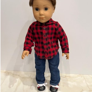 May include: A doll wearing a red and black plaid shirt, blue jeans, and black and white sneakers.