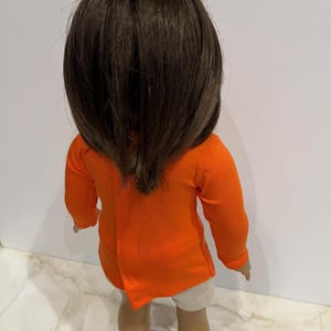 May include: A doll wearing a bright orange long-sleeved shirt and white shorts. The doll has brown hair and is wearing white shoes.