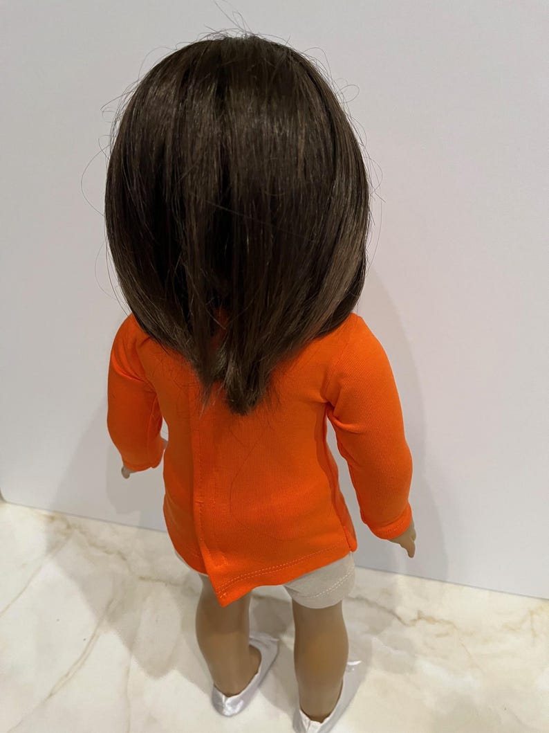 May include: A doll wearing a bright orange long-sleeved shirt and white shorts. The doll has brown hair and is wearing white shoes.