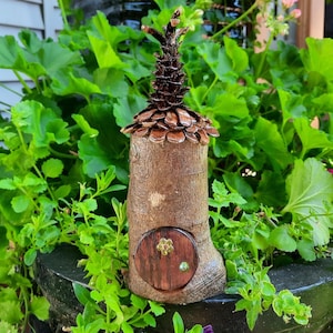 May include: A handcrafted fairy house made from a tree trunk, topped with a pinecone roof and a small wooden door. The door has a gold floral handle and a green knob. The house is set amongst vibrant green foliage.