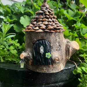 May include: A miniature fairy house crafted from a tree trunk, featuring a brown bark exterior and a roof constructed from overlapping pieces. The dark green door is complemented by a small hanging lantern. The house is set amidst green foliage.