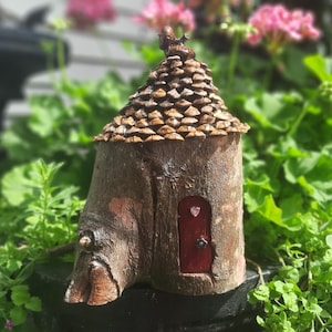 May include: A handcrafted fairy house made from a tree trunk, with a roof of pine cones and a red door with a heart. The miniature dwelling is set amidst green foliage and pink flowers, creating a charming garden scene.