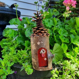 May include: A charming fairy house made from a tree trunk section, with a pine cone roof. The miniature dwelling has a small red door with a decorative key, set amidst vibrant green foliage and pink flowers.