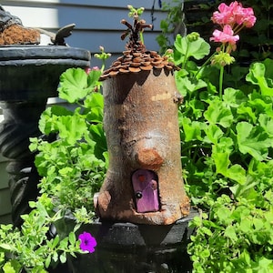 May include: A charming fairy house made from a tree trunk, with a brown bark exterior, a small purple door with a heart, and a roof of wooden pieces. It's set amidst green foliage and pink flowers, creating a magical garden scene.