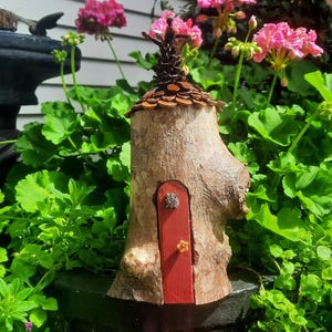 May include: A rustic fairy house crafted from a tree stump, with a red door and a roof constructed from wooden shingles. The house is set amidst vibrant green foliage and pink flowers, creating a delightful garden setting.