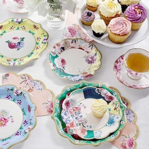 May include: A set of floral patterned paper plates with scalloped edges. The plates are in various colors, including blue, green, pink, and white. There are also cupcakes with colorful frosting on a white table.