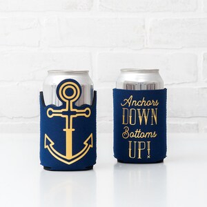 May include: Two navy blue can coolers with gold designs. One features a gold anchor, the other reads "Anchors DOWN Bottoms UP!" in gold lettering. The coolers are designed to hold standard-sized beverage cans.