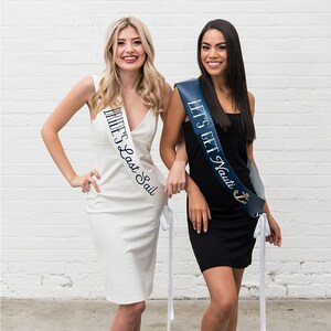 May include: Two women wearing sashes with text. One sash is white with the text "Bride's Last Sail" in navy blue. The other sash is navy blue with the text "Let's Get Nauti" and an anchor graphic in gold.