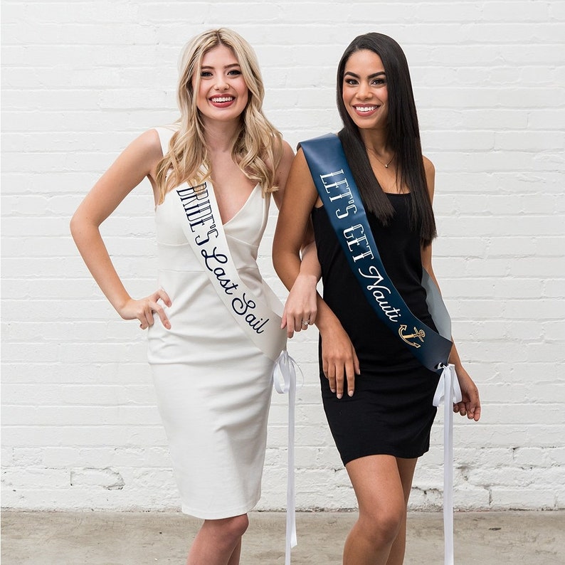 May include: Two women wearing sashes with text. One sash is white with the text "Bride's Last Sail" in navy blue. The other sash is navy blue with the text "Let's Get Nauti" and an anchor graphic in gold.
