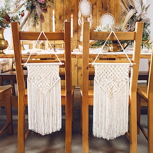 May include: Two wooden chairs with white macrame wall hangings attached to the back. The chairs are set at a wooden table with a white tablecloth.
