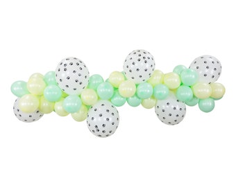 May include: A balloon garland featuring white balloons with black paw prints interspersed with pastel green and yellow balloons. The garland is arranged in a curved shape, suitable for a party or celebration.
