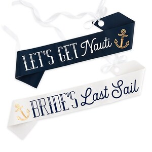 May include: Two nautical-themed sashes. One is navy blue with the text "LET'S GET Nauti" and a gold anchor. The other is white with "BRIDE'S Last Sail" and a gold anchor. Both have white ribbon ties.