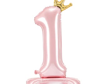 May include: Pink number one balloon with a gold crown on top. The balloon is standing on a small base.