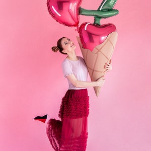 May include: A woman in a red ruffled skirt holds a large red and green balloon shaped like a cherry ice cream cone.
