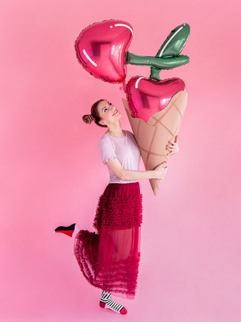 May include: A woman in a red ruffled skirt holds a large red and green balloon shaped like a cherry ice cream cone.