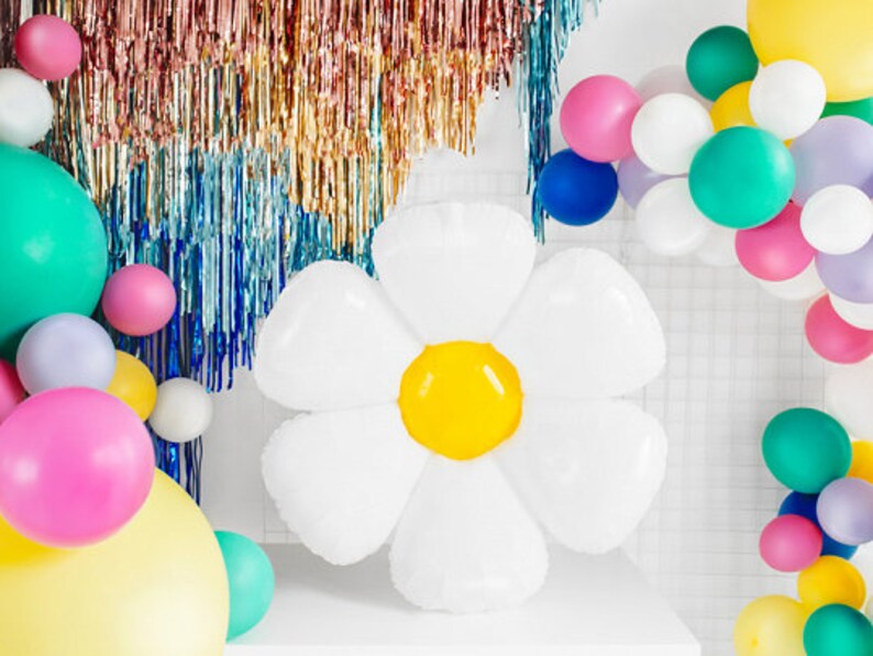May include: A large white balloon shaped like a daisy with a yellow center. The balloon is surrounded by a variety of colorful balloons, including pink, green, blue, and yellow.