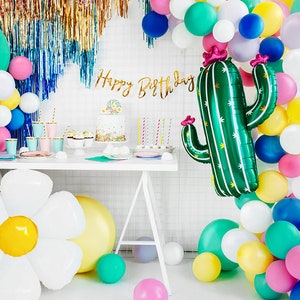 May include: A birthday party table setting with a green cactus balloon, a white daisy balloon, and a balloon arch in various colors, including pink, yellow, blue, and green. The table is decorated with a cake, cupcakes, and other party supplies. The banner says "Happy Birthday".