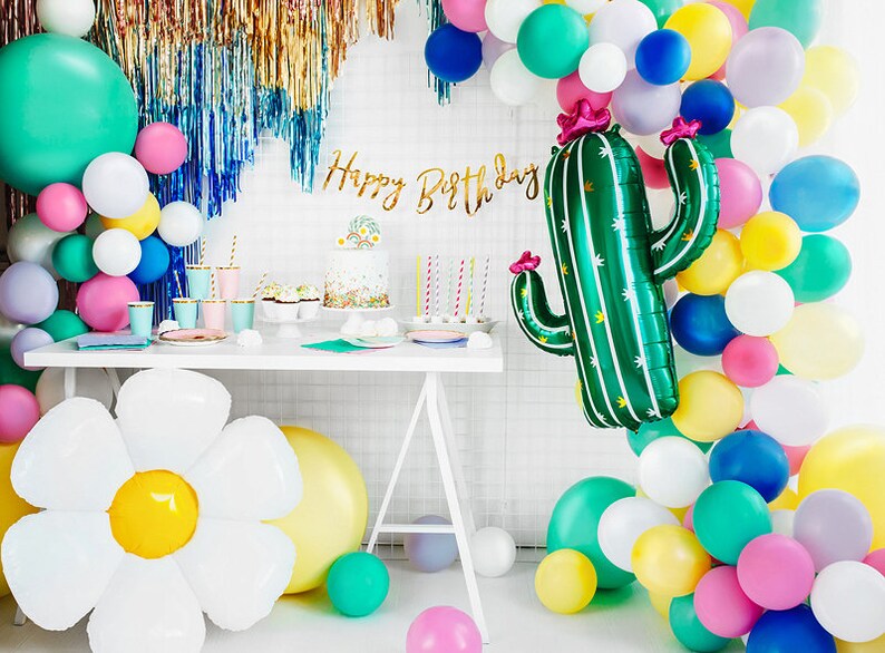May include: A birthday party table setting with a green cactus balloon, a white daisy balloon, and a balloon arch in various colors, including pink, yellow, blue, and green. The table is decorated with a cake, cupcakes, and other party supplies. The banner says "Happy Birthday".