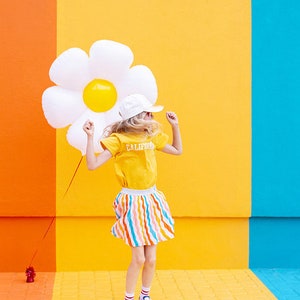 May include: A young person wearing a yellow shirt with "CALIFORNIA" printed on it, a white hat, and a colorful striped skirt, is jumping in front of a wall with orange, yellow, and blue sections. They are holding a large white inflatable daisy.