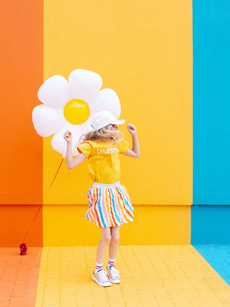 May include: A young person wearing a yellow shirt with "CALIFORNIA" printed on it, a white hat, and a colorful striped skirt, is jumping in front of a wall with orange, yellow, and blue sections. They are holding a large white inflatable daisy.