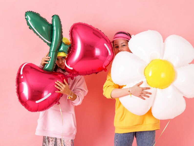 May include: Two children holding large foil balloons. One child is holding a red cherry balloon and the other is holding a white daisy balloon.