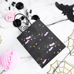 May include: Black paper gift bag with pink bats and gold stars on a white marble background. The bag is filled with black licorice, pink and white candy, and a pink honeycomb ball. There are also gold spiderwebs on the background.