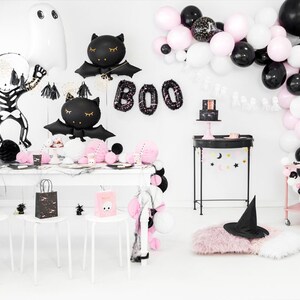 May include: A Halloween party table setting with black and pink decorations. The table is decorated with black and white balloons, a black and white skeleton, and black and white bats. The table is set with black and white plates, cups, and napkins. There is a black and white cake on the table. The table is also decorated with a black and white banner that says "BOO".