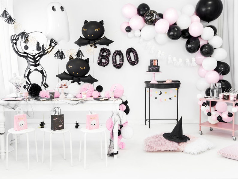 May include: A Halloween party table setting with black and pink decorations. The table is decorated with black and white balloons, a black and white skeleton, and black and white bats. The table is set with black and white plates, cups, and napkins. There is a black and white cake on the table. The table is also decorated with a black and white banner that says "BOO".