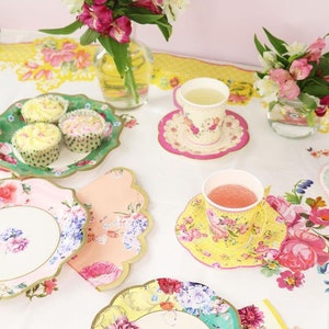May include: A floral themed tea party setting with pastel pink, yellow, and green paper plates and cups. There are cupcakes on a green plate and a bouquet of pink and white flowers in a glass vase.