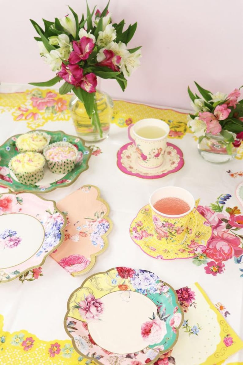 May include: A floral themed tea party setting with pastel pink, yellow, and green paper plates and cups. There are cupcakes on a green plate and a bouquet of pink and white flowers in a glass vase.